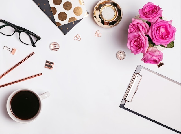 Posto di lavoro elegante con caffè, rose rosa e accessori eleganti su sfondo bianco