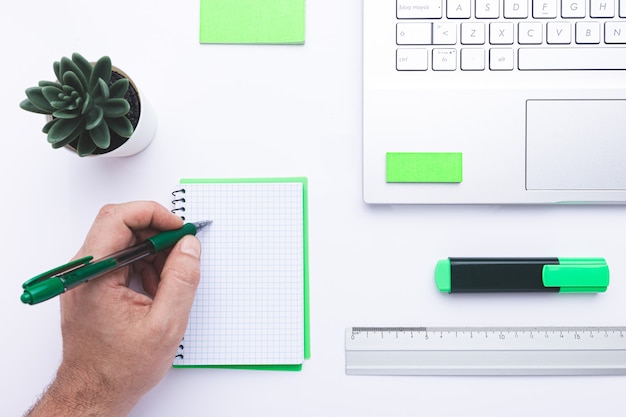 Posto di lavoro in ufficio con la scrittura a mano in notebook, laptop e articoli per ufficio verde su una scrivania bianca.