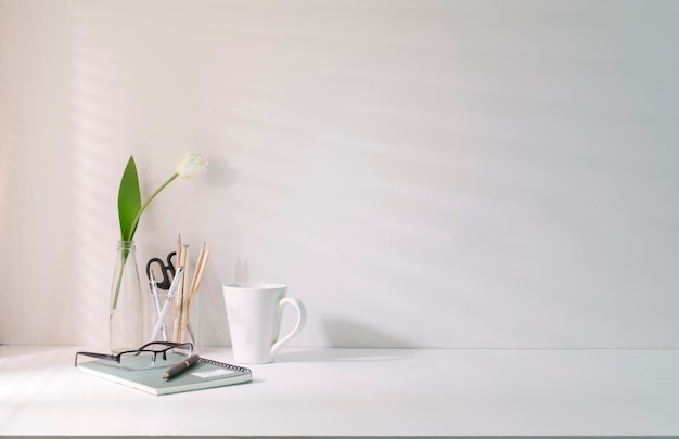 Posto di lavoro minimo con taccuino portamatite per tazza di caffè e vaso di fiori su tavolo bianco Copia spazio per il testo pubblicitario