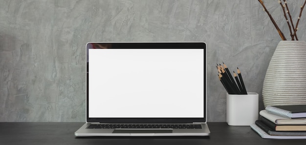 Posto di lavoro moderno con il computer portatile e gli articoli per ufficio aperti dello schermo in bianco sulla tavola nera e sulla parete grigia