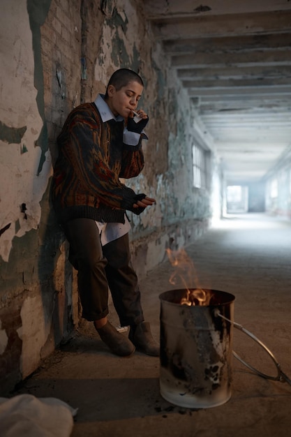 Povera giovane donna senza tetto dell'adolescente che fuma mentre si riscalda al falò in botte sopra la scena urbana abbandonata della casa del rifugio