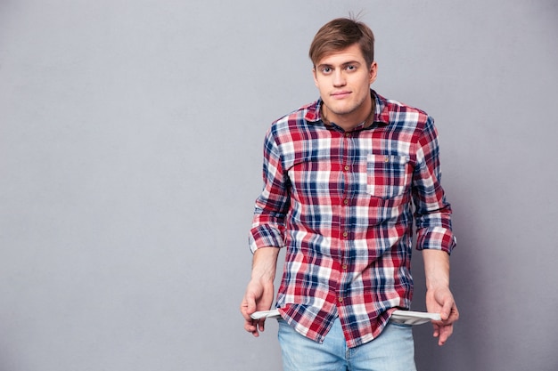 Povero bel giovane in camicia a scacchi e jeans che mostrano tasche vuote sul muro grigio