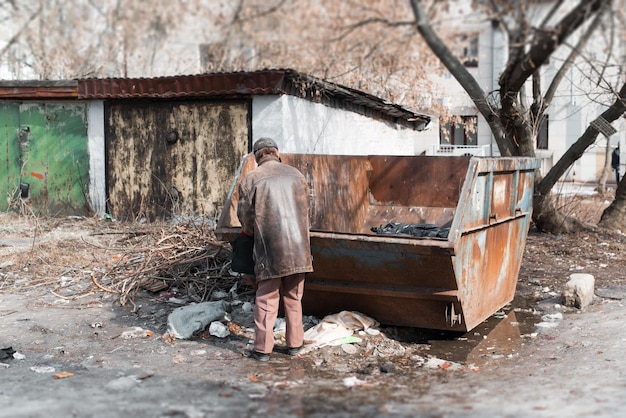 Povero senzatetto che cerca rovistando nella spazzatura in cerca di cibo e vestiti vista posteriore Povertà miseria problemi sociali concetto