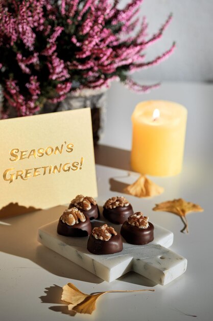 Praline di cioccolato con noce su tagliere di marmo. Biglietto di auguri con testo auguri di stagione. Disposizione autunnale - candela, foglie di ginkgo secche. Luce solare naturale, ombre. Fiori di erica rosa.