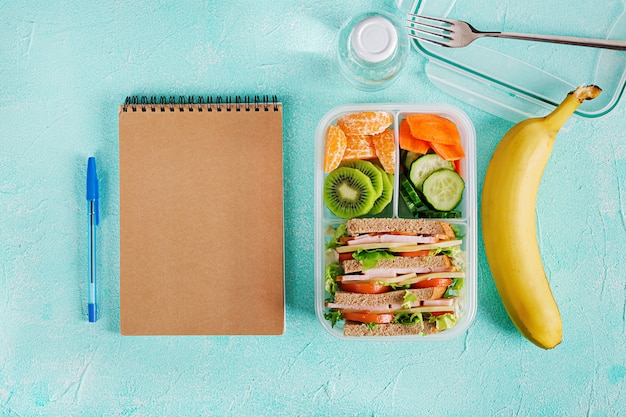Pranzo al sacco con panino, verdure, acqua e frutta sul tavolo.