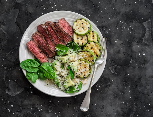 Pranzo equilibrato bistecca di manzo arrosto medio zucchine grigliate e couscous insalata di spinaci avocado su sfondo scuro vista dall'alto