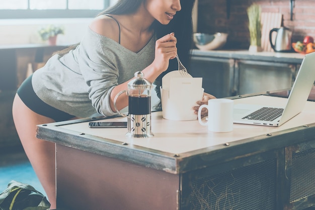 Pranzo veloce. Primo piano di una bellissima giovane donna di razza mista che mangia cibo dal contenitore e guarda il laptop mentre si appoggia alla scrivania della cucina a casa