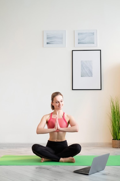 Pratica yoga Donna sportiva Allenamento online Mente armoniosa Signora atletica sorridente in abbigliamento sportivo in posa di loto Mani di namaste davanti al computer portatile nell'interno della stanza luminosa