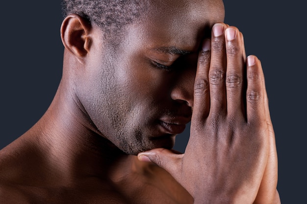 Pregando l'uomo. Vista laterale di un giovane africano che prega tenendo le mani giunte vicino e contro uno sfondo grigio
