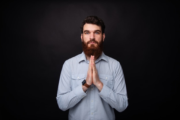 Preghiamo! Il giovane uomo barbuto si tiene per mano in posa di preghiera su sfondo nero.
