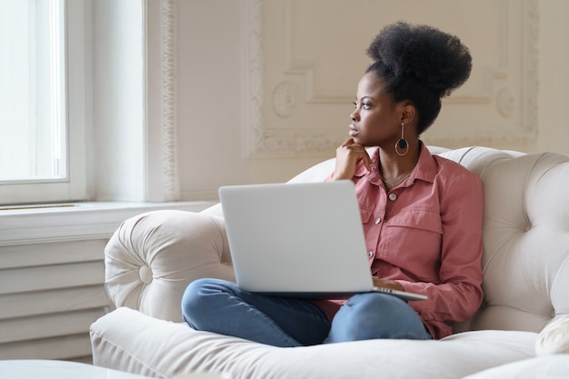 Premurosa donna africana seduta sul divano a casa, distratta dal lavoro del laptop guardando fuori dalla finestra