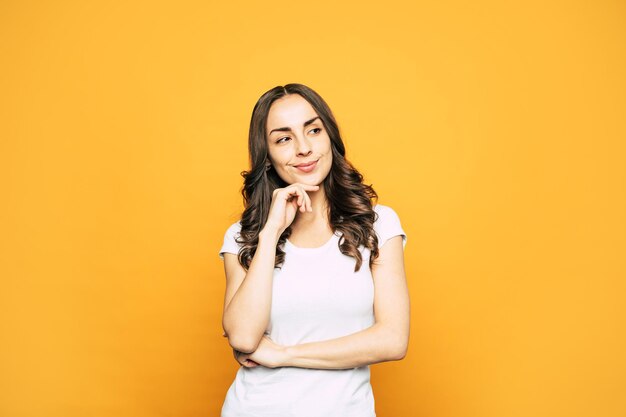 Premurosa giovane donna in maglietta bianca casual con capelli color cioccolato fondente, leggero sorriso chirk e sguardo malinconico che racconta il processo di pensiero