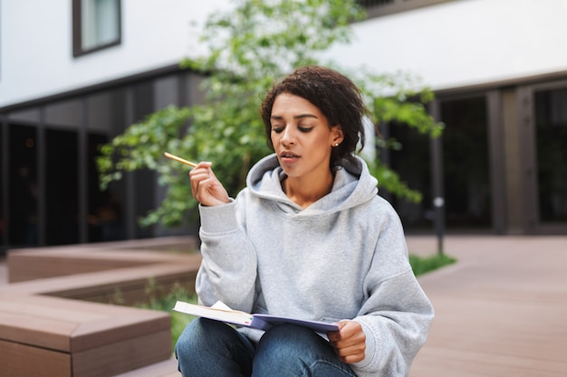 Premurosa signora con i capelli ricci scuri che si siede con il taccuino sulle ginocchia e studia da solo mentre trascorre il tempo nel cortile dell'università