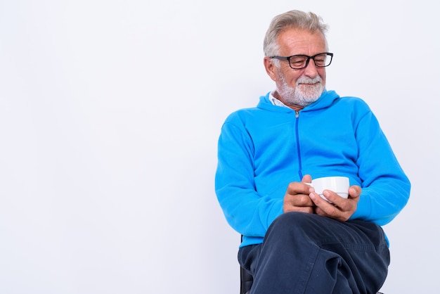 premuroso felice senior barbuto uomo sorridente mentre si tiene la tazza di caffè e seduto su una sedia pronto per la palestra su bianco