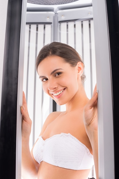 Prendere il sole in solarium. Attraente giovane donna in piedi nel solarium verticale e sorride alla telecamera