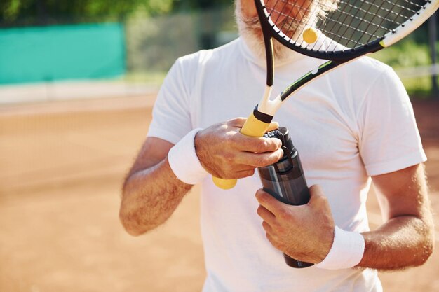 Prendersi una pausa e bere acqua Uomo anziano moderno ed elegante con racchetta all'aperto sul campo da tennis durante il giorno