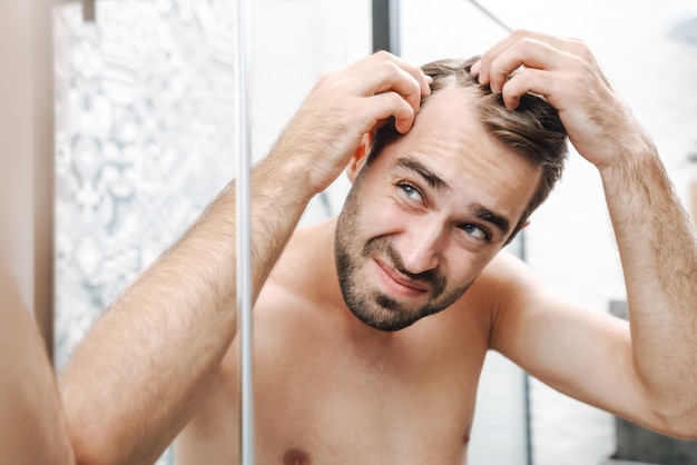 Preoccupato giovane uomo a torso nudo che si esamina i capelli mentre guarda lo specchio del bagno