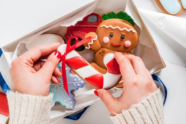 Preparando il Natale, decorando il tradizionale pan di zenzero con glassa di zucchero multicolore, la ragazza piega i biscotti in una confezione regalo bianca, con fiocco in nastro, su un tavolo di marmo bianco copyspace vista dall'alto