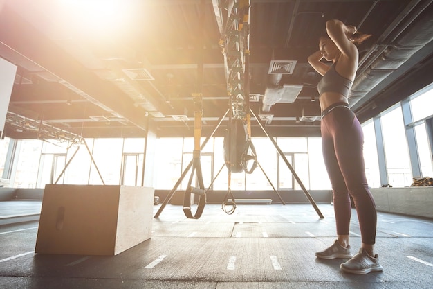 Preparazione per l'allenamento. Vista laterale di una forte donna atletica in abbigliamento sportivo che regola i capelli mentre si trova in una palestra industriale, esercitandosi con cinghie fitness trx Sport e stile di vita sano