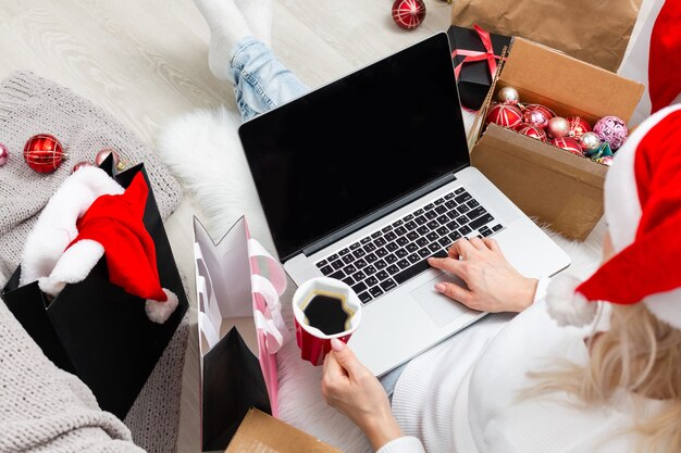 Preparazione per la festa di Natale. Donna che ordina regali e decorazioni su laptop, seduta tra scatole regalo e pacchetti, spazio copia