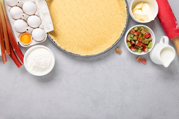 Preparazione torta crostata, pasta ricetta con uovo, panna, farina, burro, rabarbaro e mattarello su sfondo grigio. Copia spazio, vista dall'alto