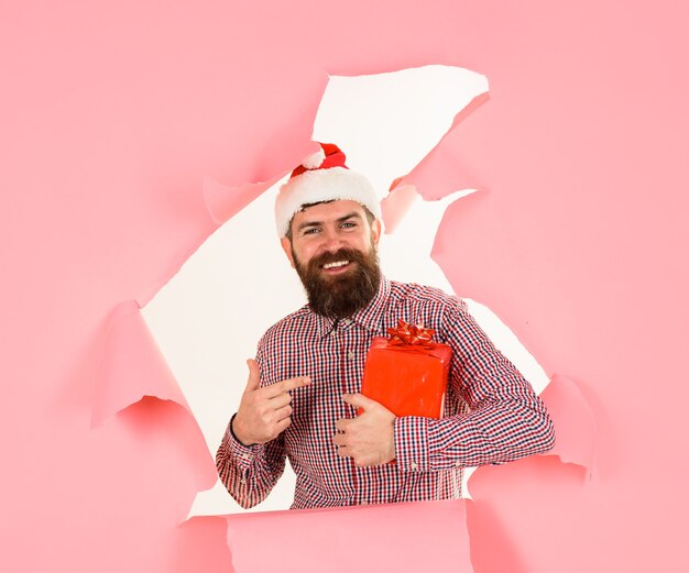 Presenta confezione regalo Babbo Natale tiene regalo uomo barbuto in cappello di Babbo Natale mostra con il dito sulla confezione regalo