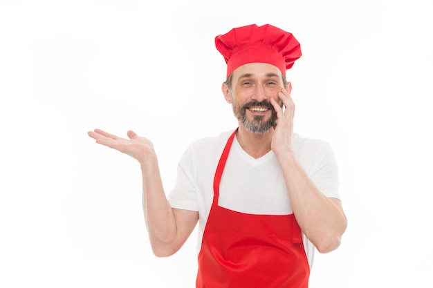 Presentazione del prodotto. Uomo maturo barbuto in cappello da chef e grembiule che indica la mano da parte. Cuoco senior con barba e baffi che indossa un grembiule con pettorina. Capo cuoco maturo in grembiule da cucina rosso, copia dello spazio.