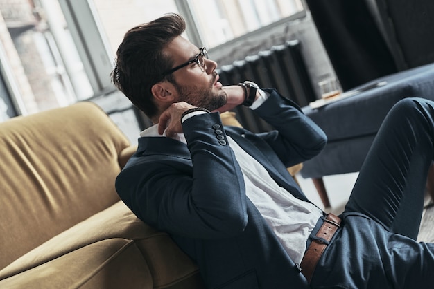 Prestando attenzione ad ogni dettaglio. Bel giovane in completo che si aggiusta la camicia mentre è seduto per terra a casa