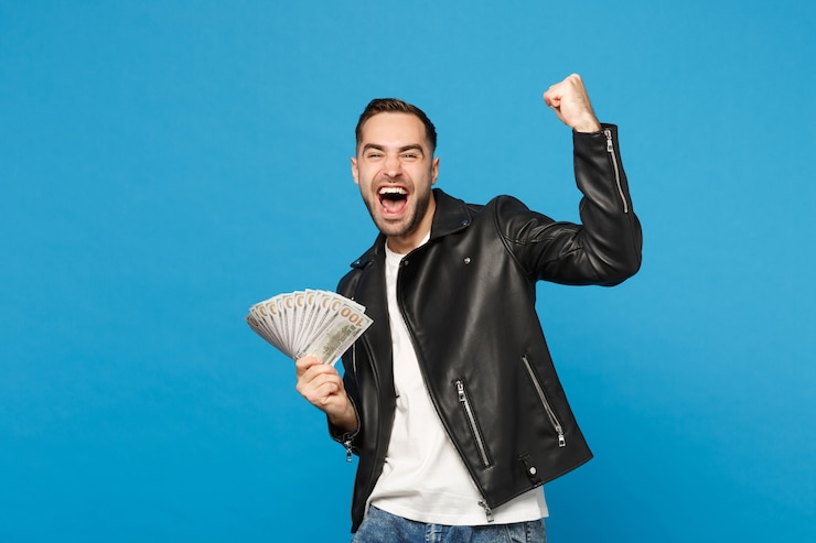 Photo pria muda belum bercukur mengenakan jaket kulit hitam dan t-shirt putih memegang setumpuk uang tunai berupa uang kertas dolar yang diisolasi dengan latar belakang dinding biru di studio. konsep gaya hidup orang. contoh ruang kosong.