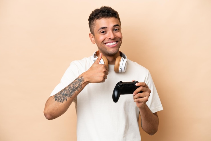 Photo pria muda brasil bermain dengan pengontrol video game terisolasi di latar belakang krem memberikan isyarat jempol ke atas