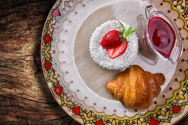 Prima colazione. Croissant con ricotta, fragole e marmellata, su un piatto colorato, su una tavola di legno. Vista dall'alto