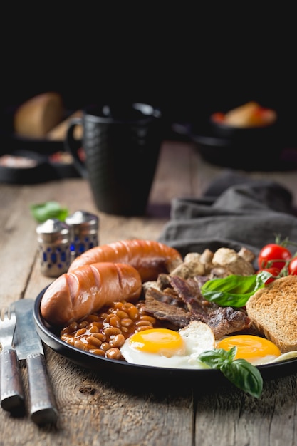 Prima colazione inglese completa con uova fritte, salsicce, pancetta, fagioli, pane tostato e pomodori su legno rustico