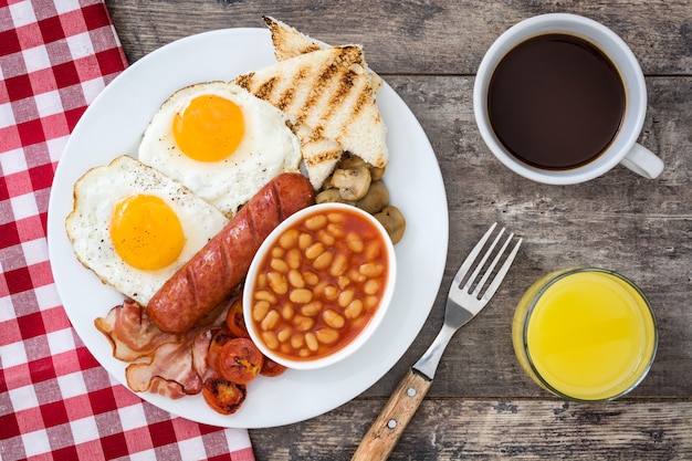 Prima colazione inglese completa tradizionale con le uova fritte, le salsiccie, i fagioli, i funghi, i pomodori arrostiti e la pancetta affumicata sulla vista superiore della superficie di legno