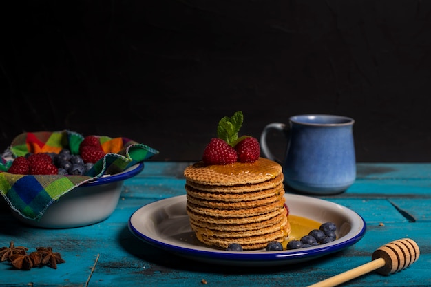 Prima colazione olandese della cialda con il lampone, il mirtillo e il miele con le foglie dell'anice su fondo di legno blu. Immagine isolata Immagine frontale. Concetto di colazione.