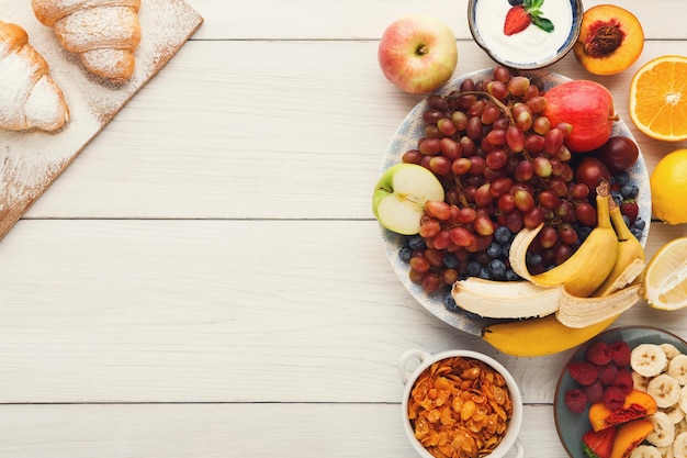 Prima colazione sana fresca con lo spazio della copia della frutta. Ciotola con uva, frutti di bosco e mela con muesli, yogurt e croissant freschi sul tavolo di legno, vista dall'alto