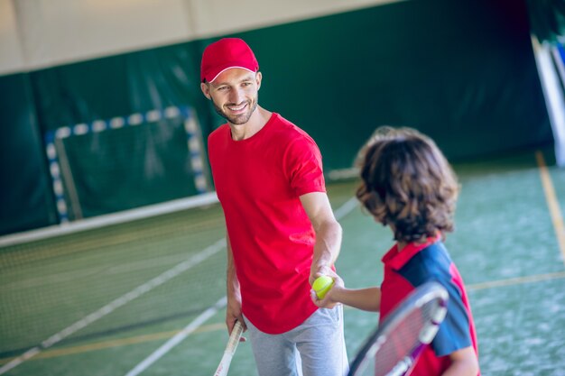 Prima della partita. Giovane uomo barbuto in un berretto rosso che stringe la mano a un ragazzo dai capelli scuri prima di giocare a tennis