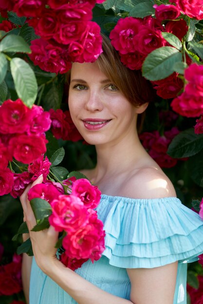 Primavera. Chiuda sul ritratto di una ragazza felice bella rossa con un sorriso affascinante che indossa un abito alla moda luce blu su una natura di rose in fiore. All'aperto.