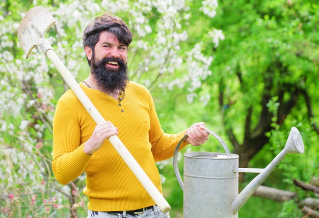 Primavera, giardiniere barbuto con annaffiatoio e vanga, uomo sorridente che si prepara a piantare, contadino che lavora in giardino.