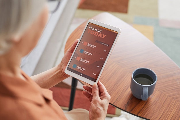 Primo piano ad alto angolo di una donna anziana moderna che utilizza tablet digitale con playlist mentre si ascolta musica a casa, copia spazio