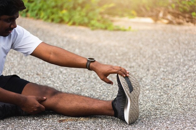 Primo piano asiatico giovane atleta sportivo corridore uomo nero indossare guarda lui seduto tirare i piedi delle dita dei piedi che allungano le gambe e il ginocchio prima di correre al parco della salute all'aperto, esercizio sano prima del concetto di allenamento