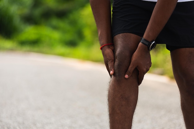 Primo piano asiatico giovane corridore sportivo uomo nero indossare orologio stand uso mani tenere comune dolore al ginocchio durante la corsa mentre si corre al parco della salute di strada all'aperto, lesioni da esercizio sano dal concetto di allenamento