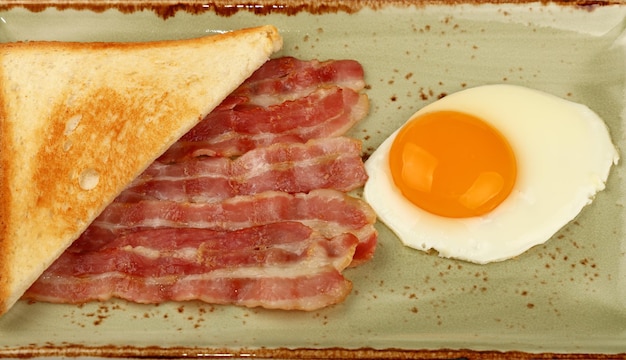 Primo piano colazione inglese, uovo lato soleggiato, metà del pane tostato e fette di pancetta arrosto sulla piastra blu, vista ad alto angolo