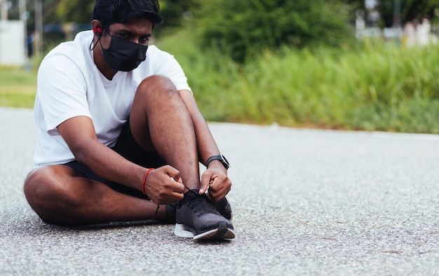 Primo piano corridore sportivo asiatico uomo nero indossare maschera protettiva lui laccio provando scarpe da corsa preparandosi per fare jogging e correre sulla strada all'aperto, concetto di allenamento esercizio fisico