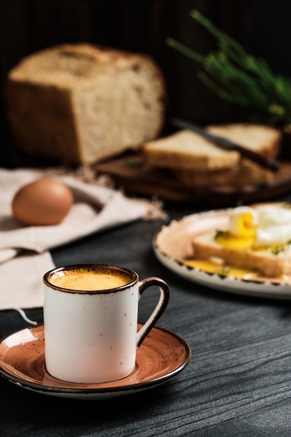 Primo piano del caffè espresso sulla tavola di legno nera. Contro il muro sfocato, uovo alla coque (in camicia) in fetta di pane, ricoperto di crema di burro ed erbe aromatiche e pagnotta di pane a fette. Idea per la colazione