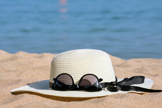 Primo piano del cappello di paglia giallo e occhiali da sole protettivi neri sulla spiaggia sabbiosa al mare tropicale in una calda giornata di sole. Concetto di vacanza estiva.