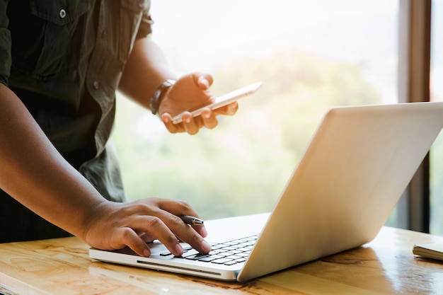 Primo piano del giovane che utilizza computer portatile e telefono cellulare nel suo ufficio che collega wifi