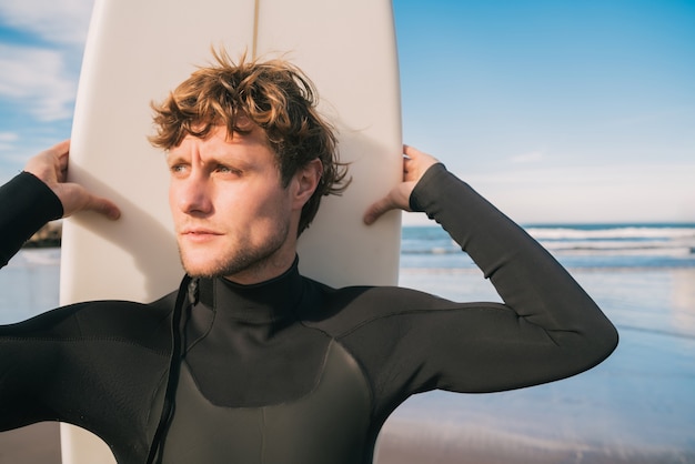 Primo piano del giovane surfista in piedi sulla spiaggia con la sua tavola da surf e indossando una tuta da surf nera