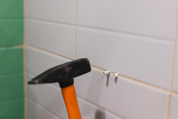 Primo piano del martello arancione che guida due tasselli nella parete piastrellata in bagno causando un forte rumore Lavori di ristrutturazione in bagno Sporco dal processo di ristrutturazione