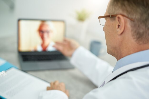 Primo piano del medico maschio seduto al tavolo con il taccuino e parlare con la paziente tramite videochiamata