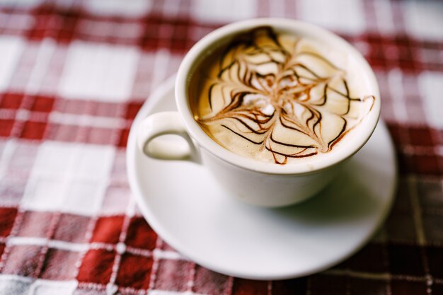 Primo piano del modello cappuccino una tazza di caffè con schiuma su un piattino su un tavolo con un rosso